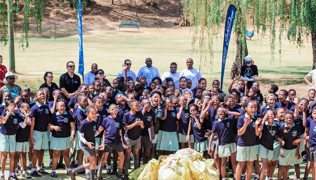 John Dory's Captain Fanplastic plastic awareness initiative. Image of large group of kids in school uniform with adults, John Dory's banners, a field, a bench, and trees in the background and yellow plastic bags piled in front.
